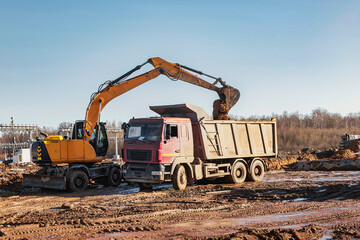 Obraz premium A wheeled excavator loads a dump truck with soil and sand. An excavator with a high-raised bucket against a cloudy sky View from the trench. Removal of soil from a construction site or quarry.
