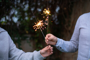 Female and male hands holding sparkling fireworks at twilight against trees in the evening. Couple celebrating holiday - 586045346
