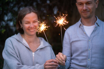Female and male hands holding sparkling fireworks at twilight against trees in the evening. Couple celebrating holiday - 586045395