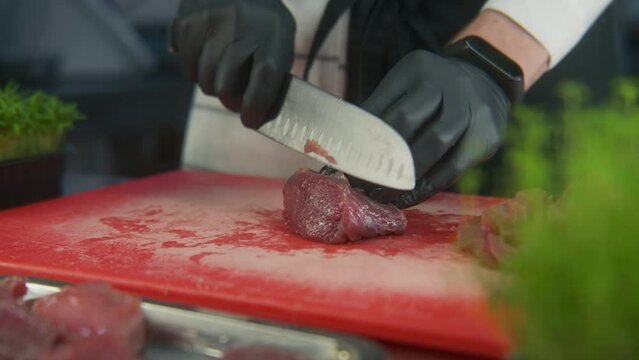 Male cook in apron cuts pieces of meat on cutting board. Boxes of microgreens stands at kitchen desk. Hands of chef close-up. Restaurant with professional cuisine. Public catering. Gastronomic art.