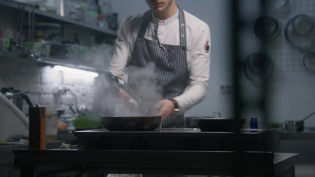 Male chef in apron sets fire to frying pan, fries beef meat in flambe style using gas burner and adds pepper. Process of preparing gourmet dish. Restaurant with professional cuisine. Gastronomic art.