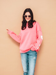 Young beautiful smiling female in trendy summer jeans and pink sweater clothes. Sexy carefree woman posing near beige wall in studio. Positive model having fun. Cheerful and happy. In sunglasses