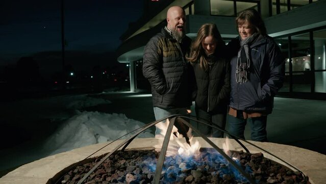Wide shot of a family huddled around an outdoor firepit at night in the winter.  The gorgeous flames flicker and create an intimate mood.  Shot in glorious 4k.