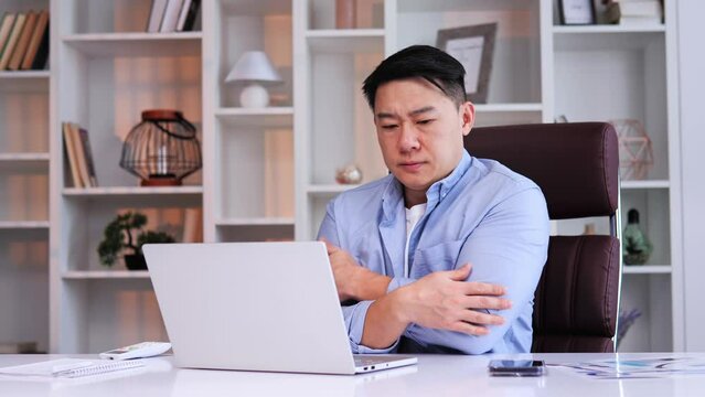 Asian Chinese office worker feels cold, rubs hands while working on laptop, turns off AC. Suffers from low indoor temp due to ventilation. Importance of temperature control in the workplace.