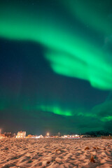 wunderbare Nordlichter über dem Dorf Hillesøy. Hell erleuchtete Häuser und Straßenlaternen bilden einen starken Kontrast zum dunklen Himmel mit der grünen Aurora Borealis. Winterstimmung in Norwegen