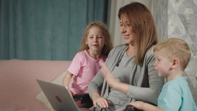 Mother with son and daughter talks on video call via laptop. Woman with paralysis in wheelchair and kids wave hello looking at screen of device closeup
