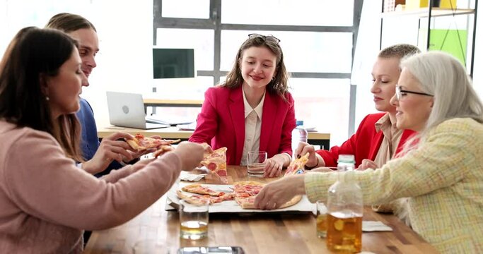 Delighted colleagues take slices of delicious pizza sitting at big wooden table in premise. Happy man and women enjoy lunch at work break in office slow motion