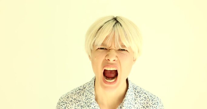 Emotional irritated blonde woman squints and open mouth screaming loud. Expressive female person shows aggression standing on beige background slow motion
