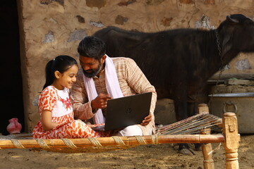 Happy Indian father and his daughter sitting together on a cot with a laptop, father teaching his daughter how to use a laptop while pointing towards the laptop.