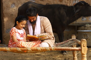 Happy rural Indian father and daughter sitting together on a cot. Father is teaching his daughter how to read with a book in his hand