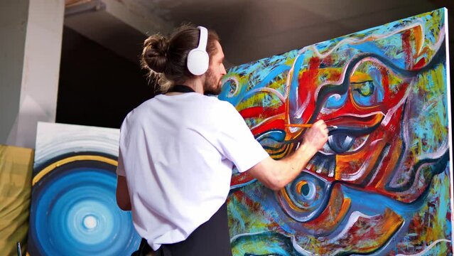Talented innovative artist in white headphones paints with brushes on a large canvas in studio, creating a colorful, emotional, and sensual oil painting. Contemporary artist creating abstract art.