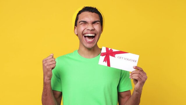 Surprised happy young man of African American ethnicity wear green t-shirt hat point finger on gift certificate coupon voucher card for store doing winner gesture isolated on plain yellow background