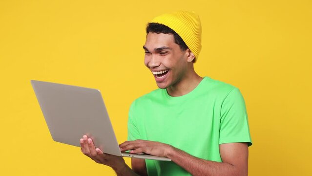 Smart IT user young man of African American ethnicity wears green t-shirt hat hold in hand use work typing on laptop pc computer isolated on plain yellow background studio. People lifestyle concept