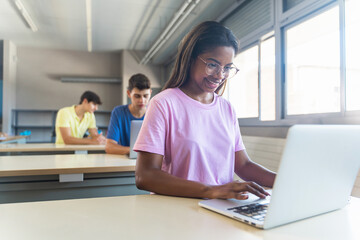 Smiling African American female student wearing eyeglasses standing using laptop in classroom