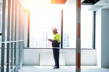Portrait of engineering man standing, looking tablet in his hand beside window at work site on building and orange of light.