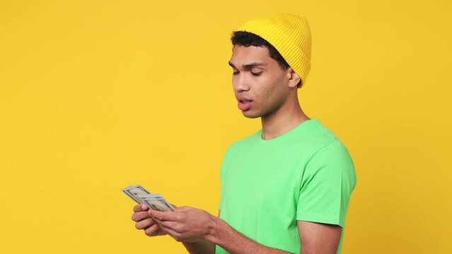 Side view happy accountant young man of African American ethnicity wear green t-shirt hat hold in hand count stack of cash money in dollar banknotes isolated on plain yellow background studio portrait