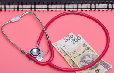 Stetoskop medyczny j polskie banknoty na różowym tle, z tyłu terminarz w formie kołonotatnika 