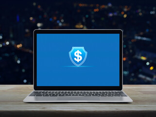 Dollar with shield flat icon on modern laptop computer monitor screen on wooden table over blur colorful night light city tower and skyscraper, Business money insurance and protection online concept