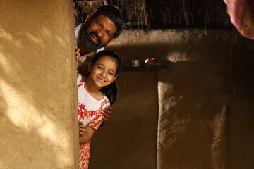 Portrait of happy rural Indian father and daughter peeking from behind the wall at the doorstep of...