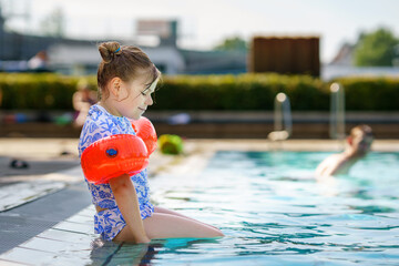 Naklejka premium Little preschool girl with protective swimmies playing in outdoor swimming pool by sunset. Child learning to swim in outdoor pool, splashing with water, laughing and having fun. Family vacations.