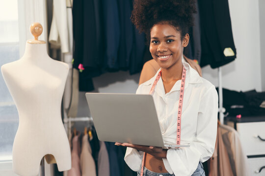 Happy beautiful African American woman fashion designer clothes working with notebook computer in own dressmaking studio or boutique	