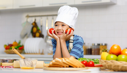 Millennial Asian little boy chef wearing tall white cook hat and apron standing holding red tomatoes near face posing in home kitchen full of fruits vegetables and sliced bread on cooking counter