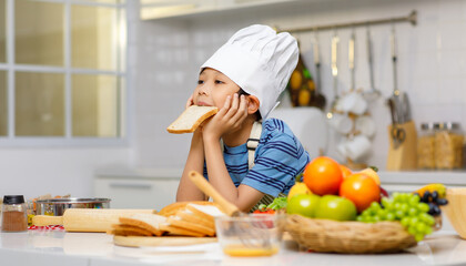 Millennial Asian little boy chef wearing tall white cook hat and apron standing posing eating sliced bread in home white kitchen full of fruits vegetables and bread on cooking counter.