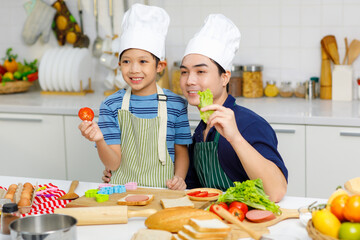 Millennial Asian little young boy chef wearing tall white cook hat and apron standing smiling holding sliced tomato posing together with father in home kitchen full of ingredients on cooking counter