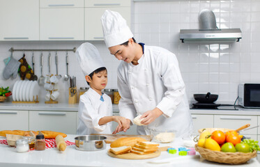 Asian young little boy chef in uniform with white tall cook hat standing learning preparing massaging flour dough on wooden cutting board while male cooking teacher helping teaching in home kitchen