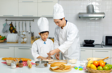 Asian young little boy chef in uniform with white tall cook hat standing learning preparing massaging flour dough on wooden cutting board while male cooking teacher helping teaching in home kitchen