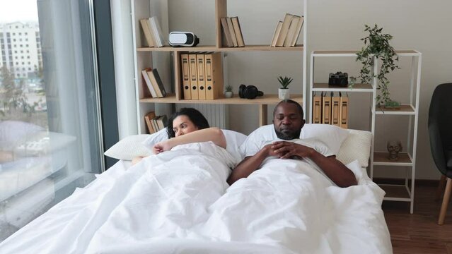 Loving african american couple in casual attire stretching their limbs after night sleep while still lying in bed of bright bedroom. Relaxed family man and woman starting new sunny day in modern flat.