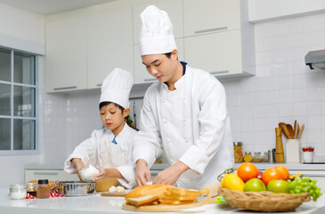 Asian young little boy chef in uniform with white tall cook hat standing learning preparing massaging flour dough on wooden cutting board while male cooking teacher helping teaching in home kitchen