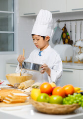 Asian professional little young boy chef in white cooking uniform and tall cook hat standing blending wet flour into wooden bowl at counter full of ingredients fruits vegetables and bread in kitchen