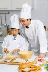 Asian young little boy pastry chef in white uniform with tall cook hat standing while male cooking teacher smiling help teaching in white  kitchen.