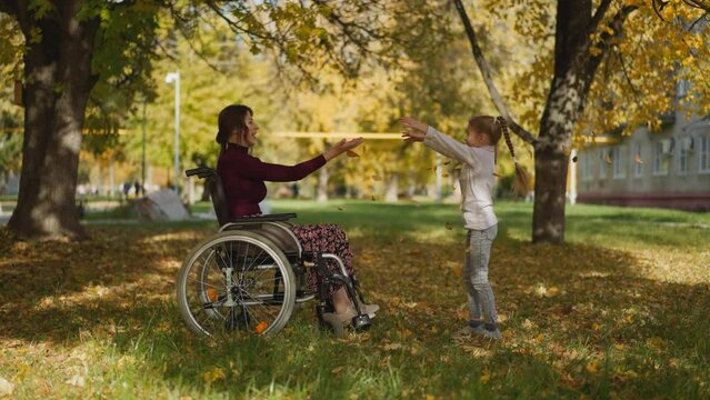 Positive adult woman with spinal cord injury in wheelchair and preschooler girl play throwing autumn leaves upward. Active family recreation during rehabilitation