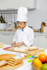 Asian professional young little boy pastry chef in white cooking uniform and tall cook hat standing using heart shape mold cutting preparing dough on wooden board at counter full of baking equipment