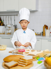 Asian professional young little boy pastry chef in white cooking uniform and tall cook hat standing using heart shape mold cutting preparing dough on wooden board at counter full of baking equipment