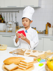 Asian professional young little boy pastry chef in white cooking uniform and tall cook hat standing using heart shape mold cutting preparing dough on wooden board at counter full of baking equipment