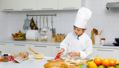 Asian professional young little boy pastry chef in white cooking uniform and tall cook hat standing using heart shape mold cutting preparing dough on wooden board at counter full of baking equipment