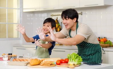 Obraz na płótnie Canvas Asian young little boy chef wearing apron standing using stainless filter sifting white flour into glass bowl while father helping at counter full of baking equipment eggs breads tomatoes in kitchen