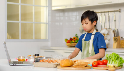 Asian young little boy pastry chef wearing apron standing kneading flour dough learning from laptop computer preparing baking bakery at counter full of equipment eggs breads vegetables in kitchen