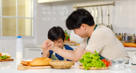 Asian young little boy chef wearing apron standing using stainless filter sifting white flour into glass bowl while father helping at counter full of baking equipment eggs breads tomatoes in kitchen