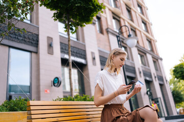 Low-angle view of charming young woman entering credit card number for online shopping using smartphone sitting on yellow bench on city street. Pretty focused female paying with card walking outdoors.