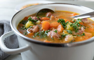 Stew with carrots, potatoes and smoked pork meat in a white enamel pot.