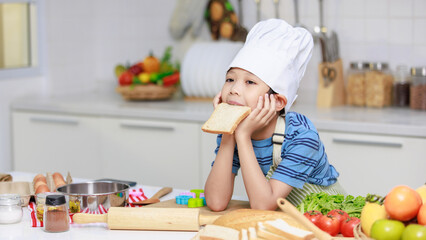 Millennial Asian little boy chef wearing tall white cook hat and apron standing posing eating...