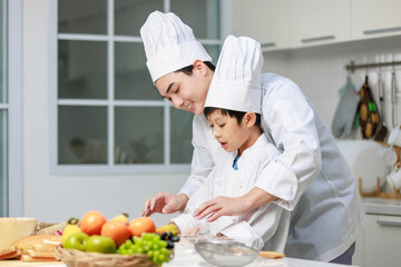 Asian young little boy chef in uniform with white tall cook hat standing learning preparing massaging flour dough on wooden cutting board while male cooking teacher helping teaching in home kitchen