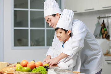 Asian young little boy chef in uniform with white tall cook hat standing learning preparing massaging flour dough on wooden cutting board while male cooking teacher helping teaching in home kitchen