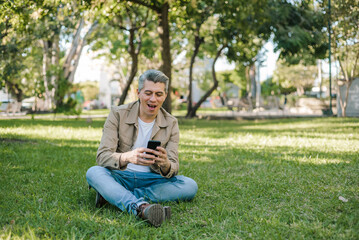 Gray-haired man looking at his cell phone sitting on the grass in a park.