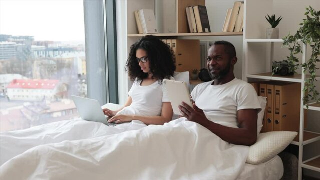 Front view of multicultural couple in love working on modern gadgets while lying under blanket at home. Smiling man reading social updates via tablet and serene woman shopping online using laptop.