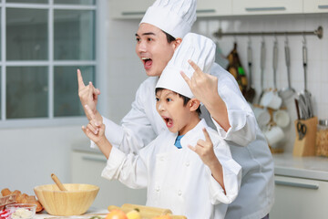 Asian young little boy chef and male cooking teacher in uniform with white tall cook hat standing holding rock star hands sign up open mouth wide screaming shouting posing together in home kitchen